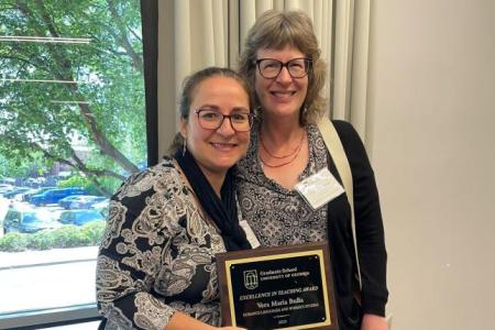 Photo of two women standing in front of a large window. The woman on the left is holding a plaque commemorating her Excellence in Teaching Award