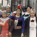 Seven Mapuche women of various ages wearing traditional clothing in shades of blue, purple, red, and black. They stand together in front of a building, posing for the camera.